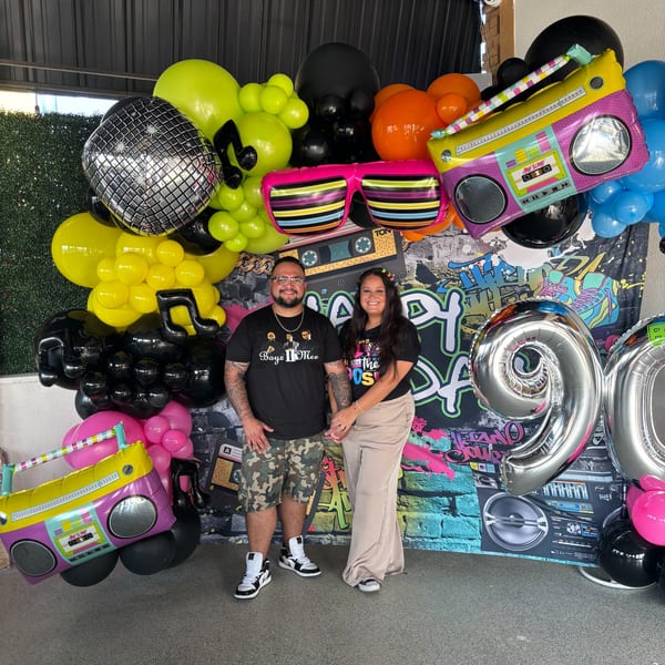 A couple stands in front of a colorful 90s-themed balloon and graffiti backdrop.
